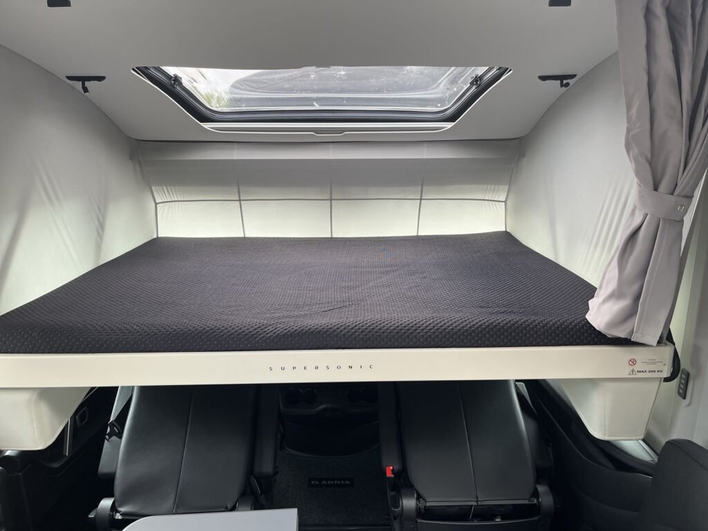 Obrázek interiéru obytného auta, výklopné lůžko a pohodlné sedadla. Vůz má značku Adria Supersonic.