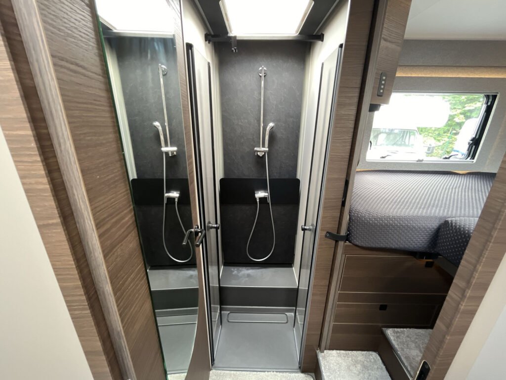 Obrázek interiéru obytného auta s koupelnou a spací zónou. Moderní design s dvoudveřovým sprchovým koutem.