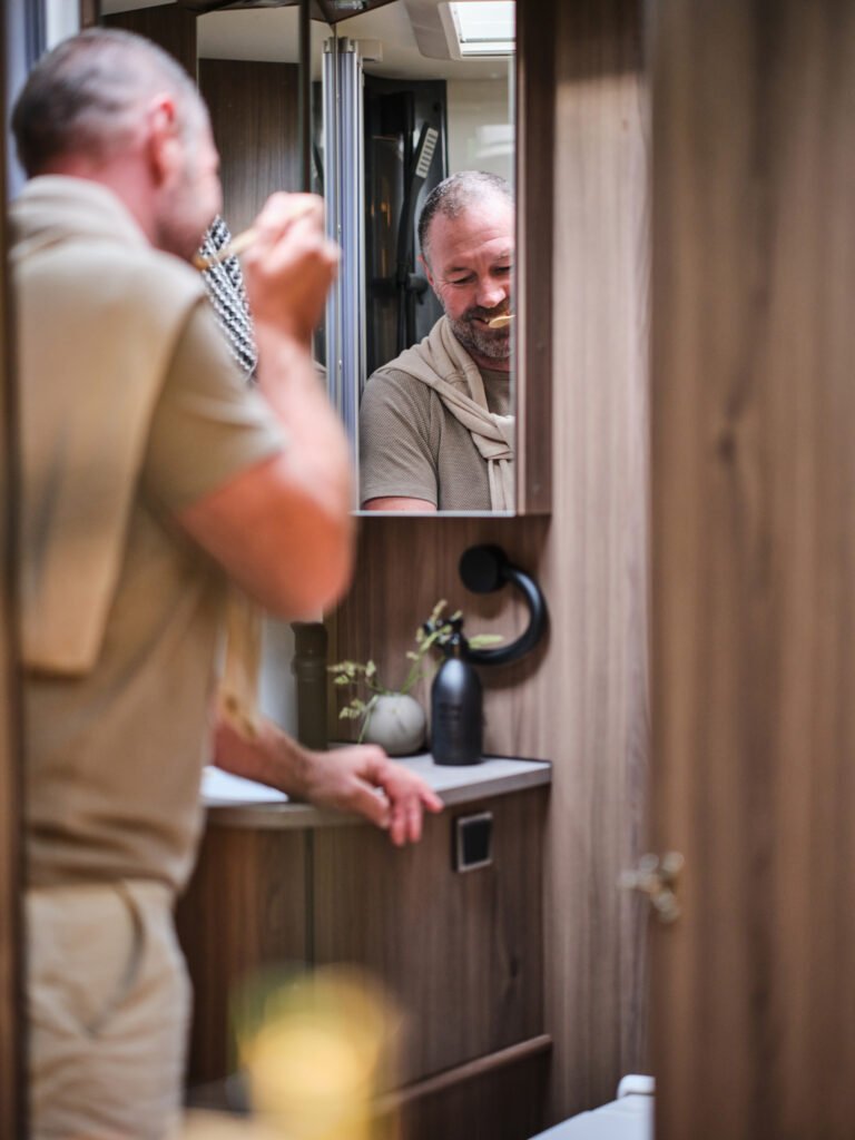 Na obrázku je interiér karavanu, muž se připravuje u zrcadla. Karavan nemá viditelnou značku ani model.