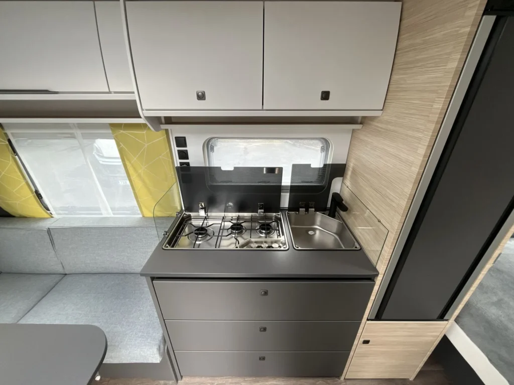 Obrázek interiéru karavanu s moderní kuchyňkou, sporákem a umyvadlem. Elegantní design pro pohodlné cestování.