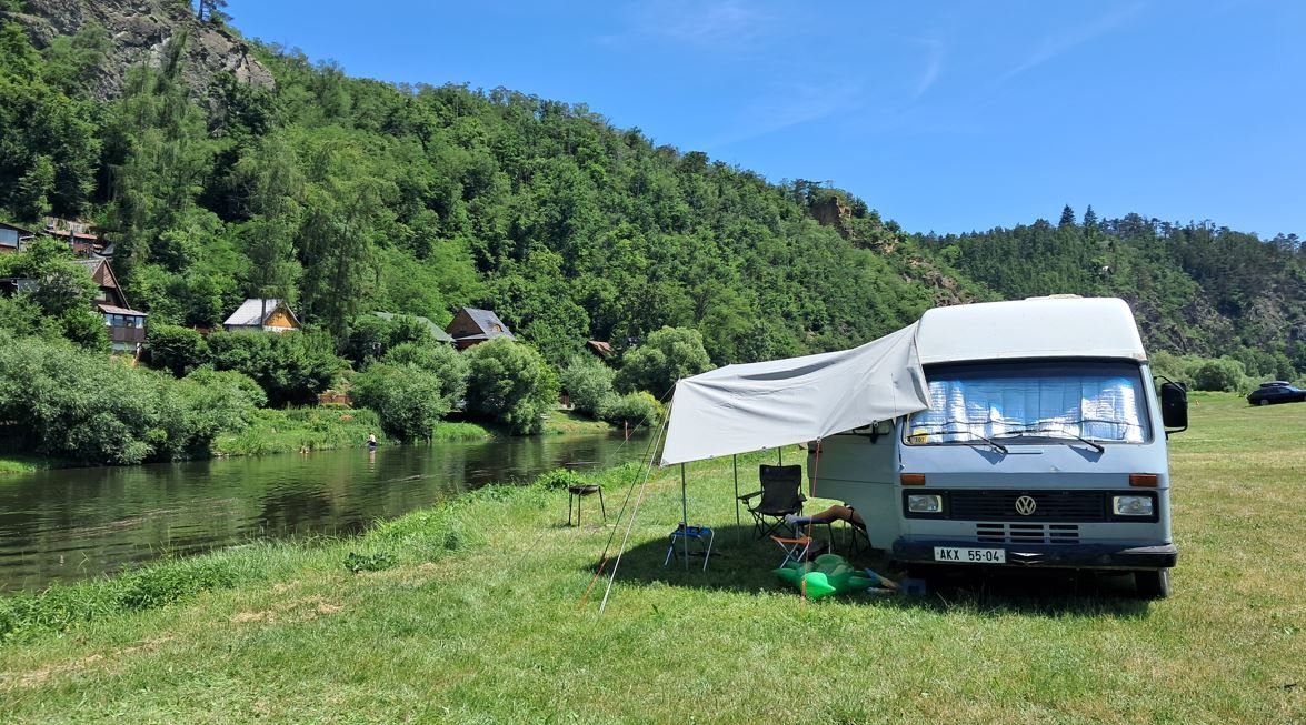 Obytné auto Volkswagen s markýzou u řeky, perfektní místo pro odpočinek v přírodě.