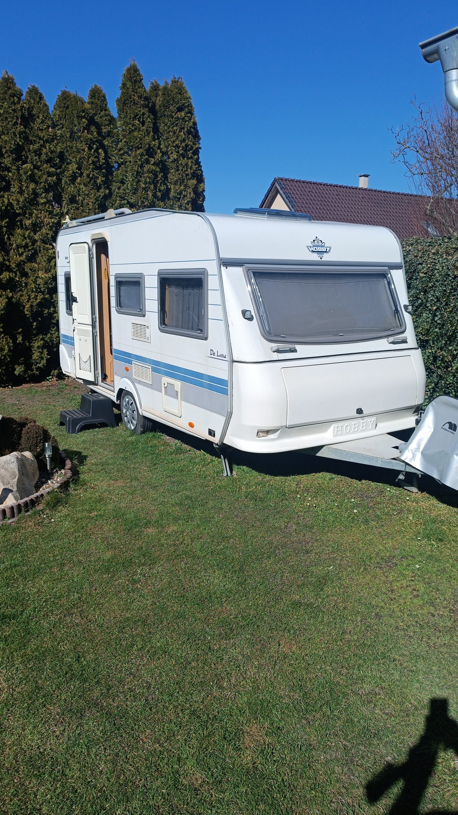 Karavan Hobby De Luxe, vystavený na trávníku, připravený na dobrodružství v přírodě.