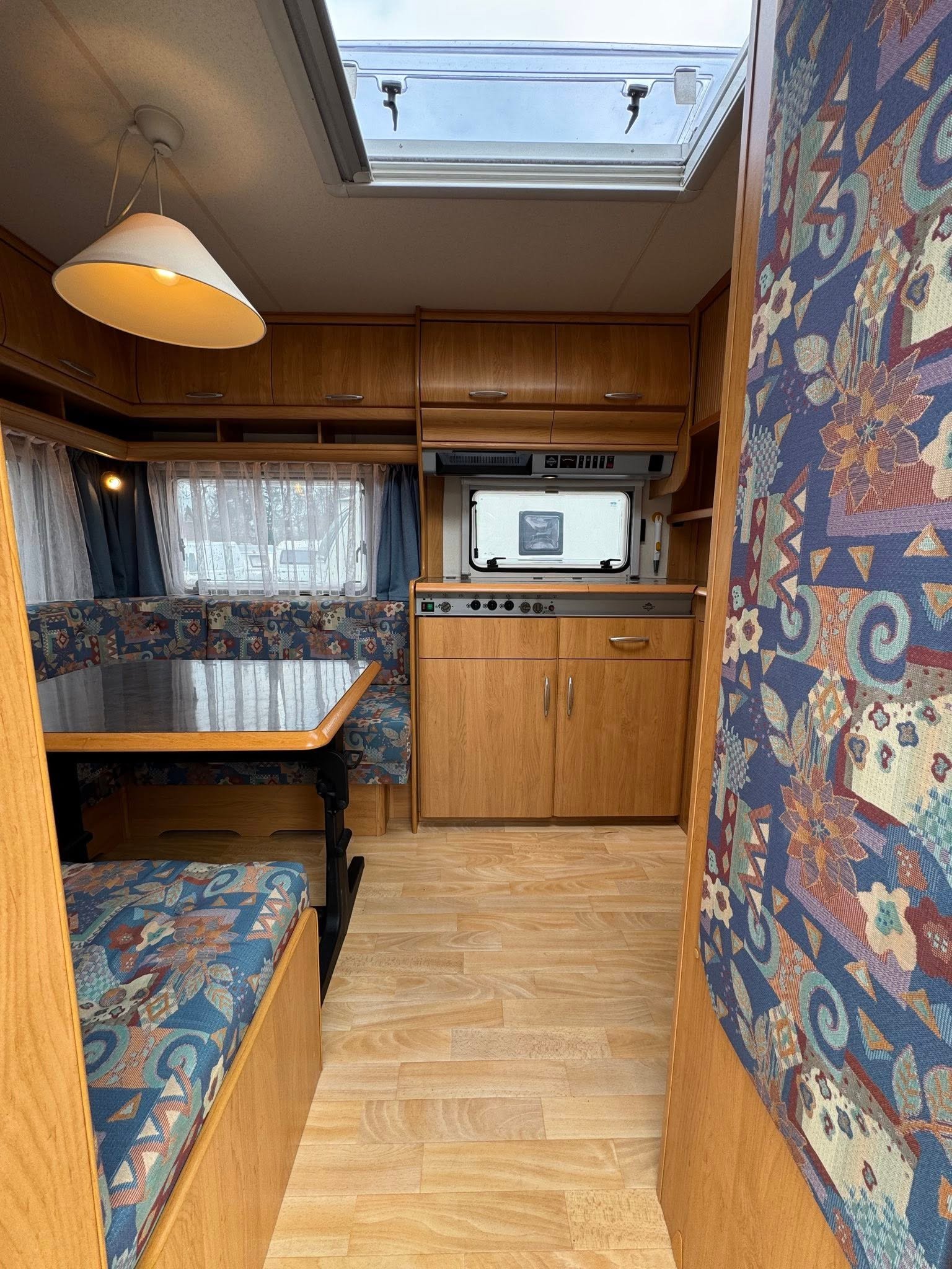 Interiér karavanu s kuchyňkou a stoly, dřevěný nábytek a barevné čalounění. Ideální pro pohodlné cestování.