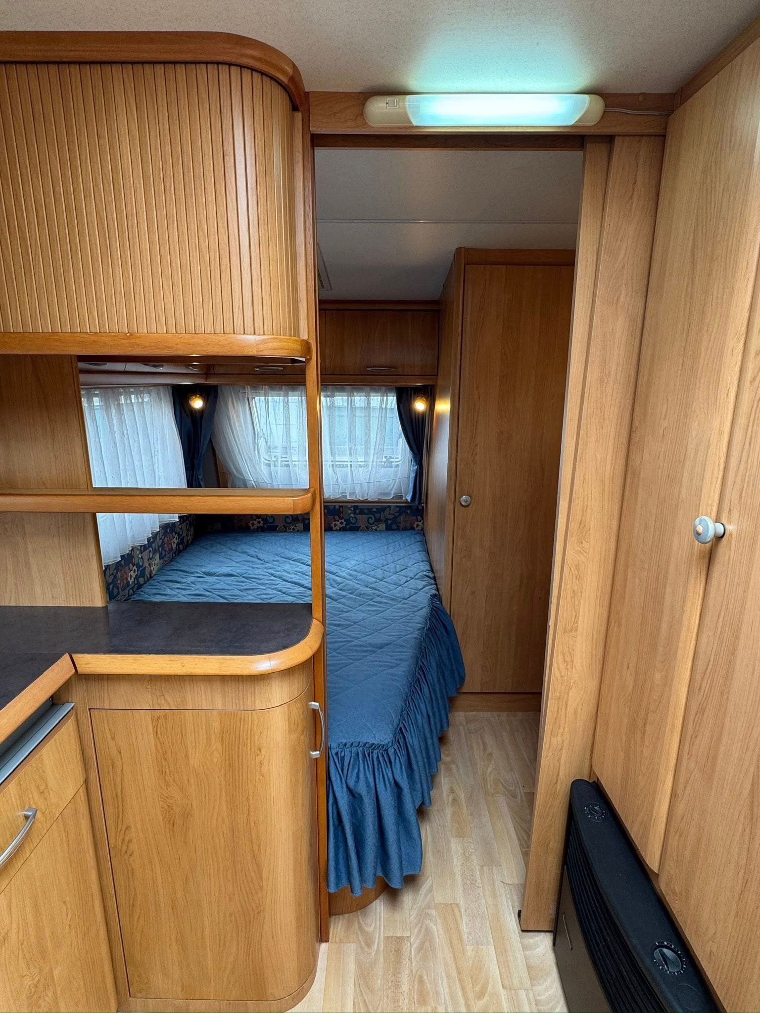 Obrázek interiéru karavanu s dřevěnou výzdobou a dvojlůžkem v modré barvě. Komfortní prostor pro cestování.