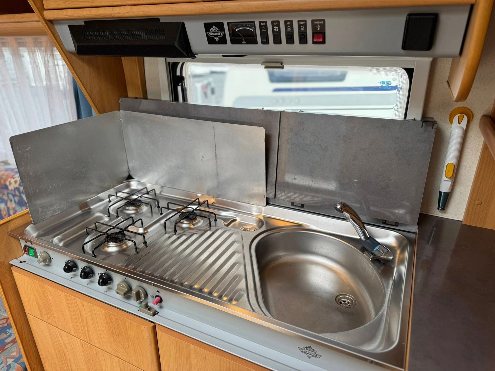 Obrázek ukazuje kuchyňku v karavanu, se sporákem a dřezem, ideální pro campingové vaření.