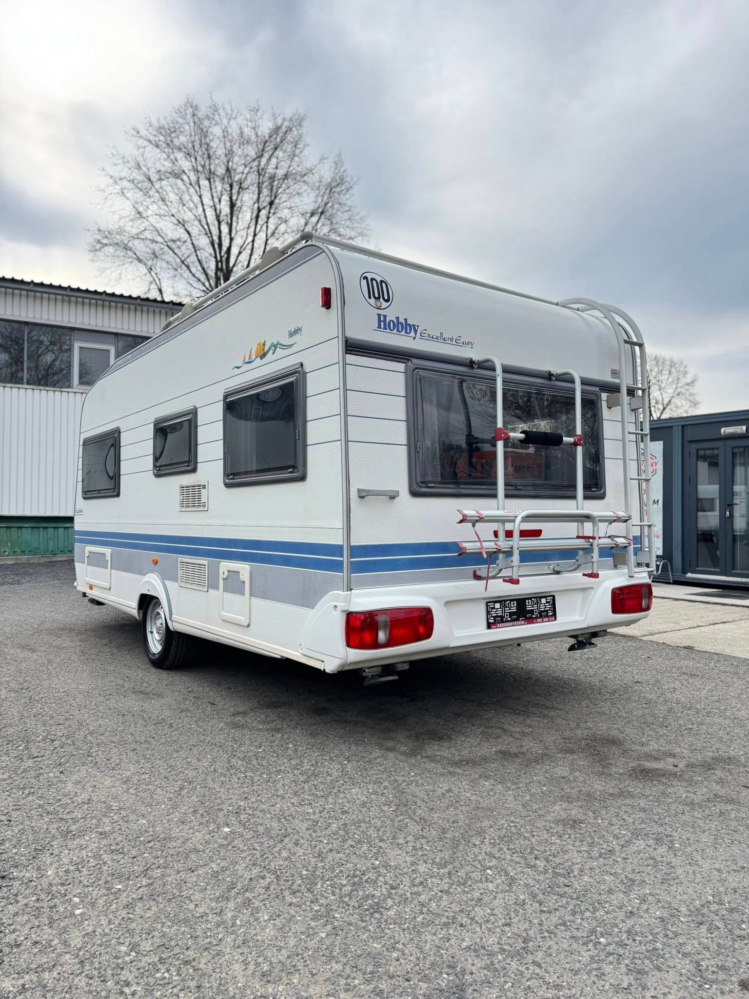 Karavan Hobby Excellent Easy s modrými pruhy a zadním žebříkem. Ideální pro kempování a dobrodružství.