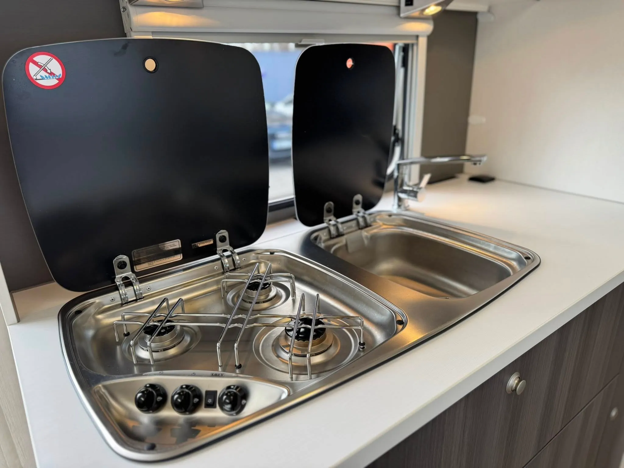 Obrázek ukazuje moderní kuchyňku s vařičem a dřezem, ideální pro obytné auto nebo karavan. Bez značky či modelu.