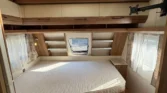 Obrázek zobrazuje interiér karavanu s manželskou postelí a praktickým úložným prostorem.