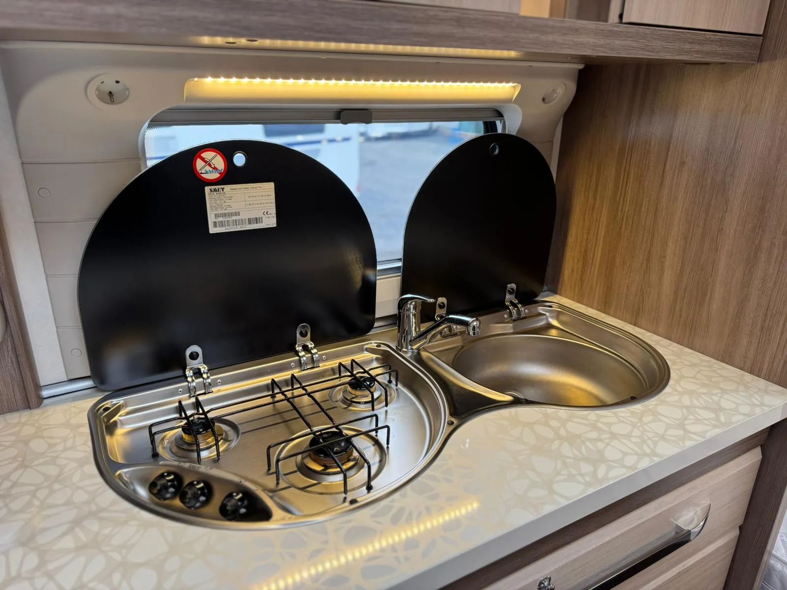 Obrázek zobrazuje kuchyňský kout obytného auta s varnou deskou a dřezem. Ideální pro mobilní vaření.