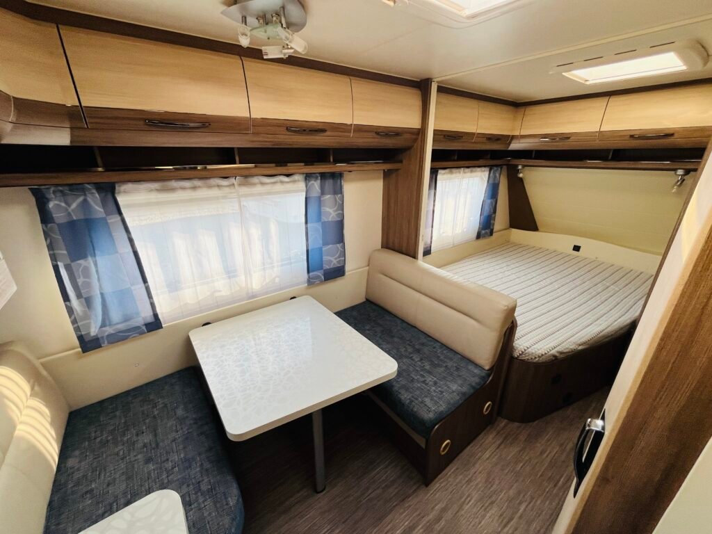 Obrázek interiéru karavanu, moderní design s dvojlůžkem a jídelním stolem. Ideální pro rodinné výlety.