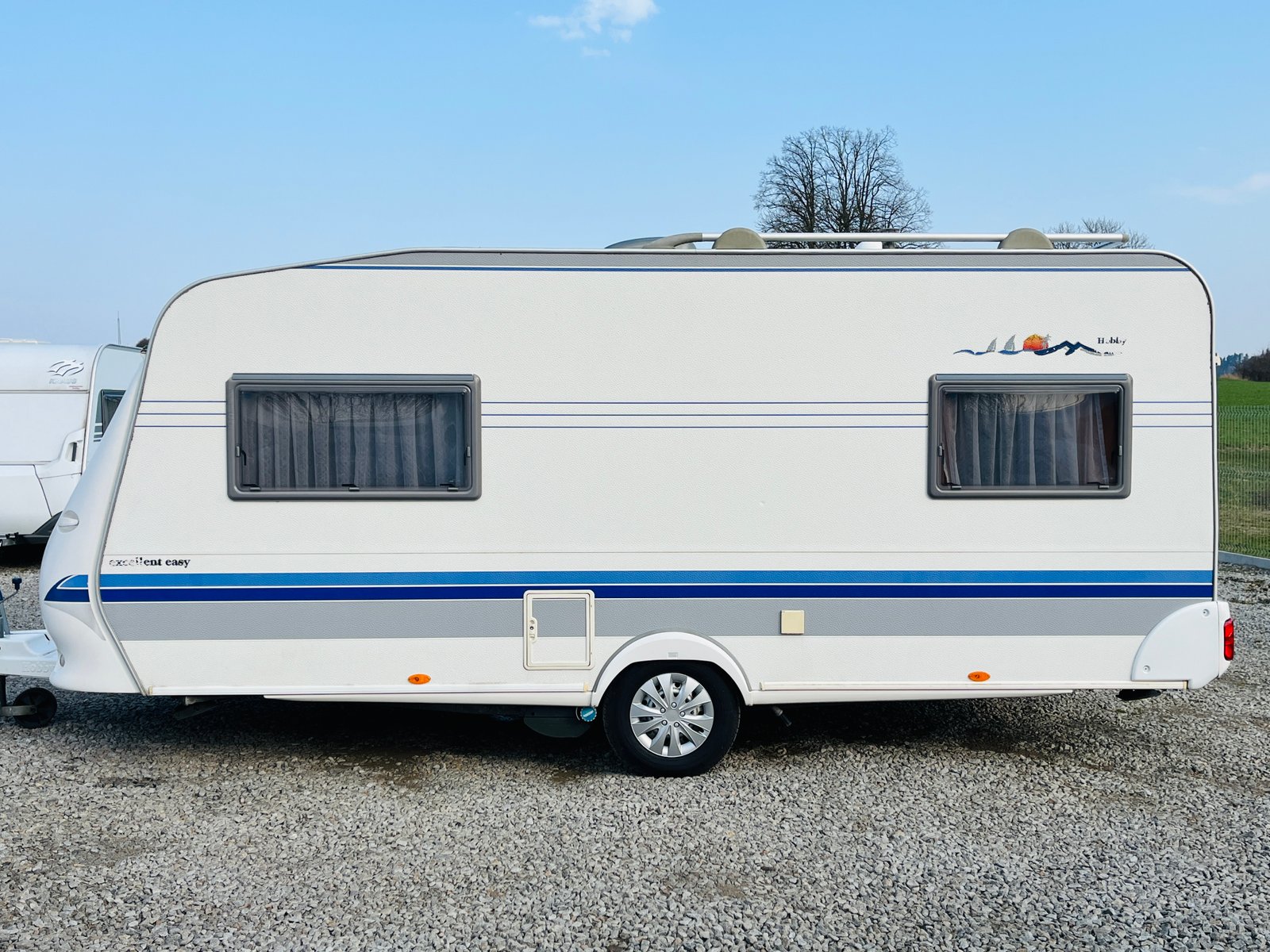 Karavan Hobby Excellent Easy, bílý s modrými pruhy, ideální pro pohodlné cestování.