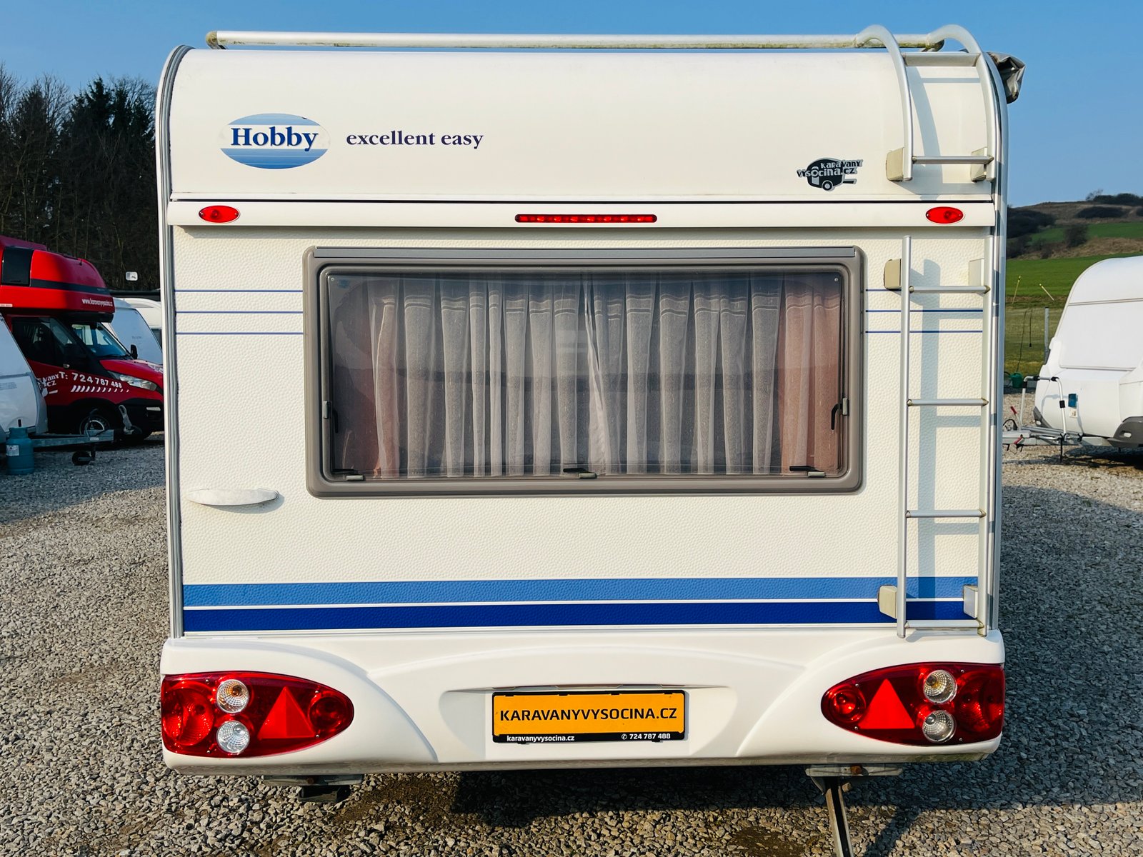 Karavan Hobby Excellent Easy s bílou karoserií a modrými pruhy, ideální pro pohodlné cestování.