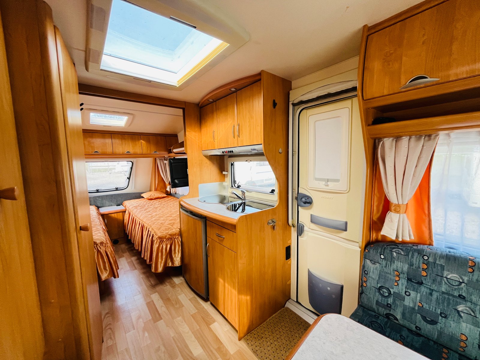 Interiér karavanu s dřevěným nábytkem a kuchyňským koutem. Obytný prostor s postelemi a útulným posezením.