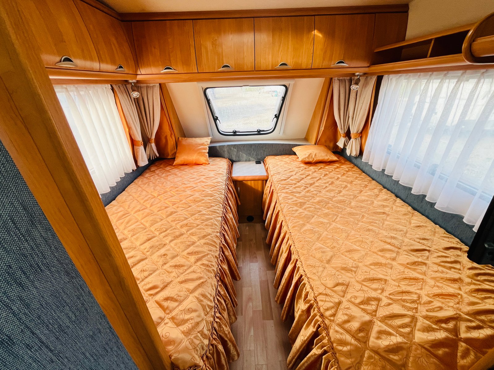 Obrázek interiéru karavanu s dvojlůžkem a dřevěnými skříňkami. Dominantní oranžové povlečení dodává teplou atmosféru.