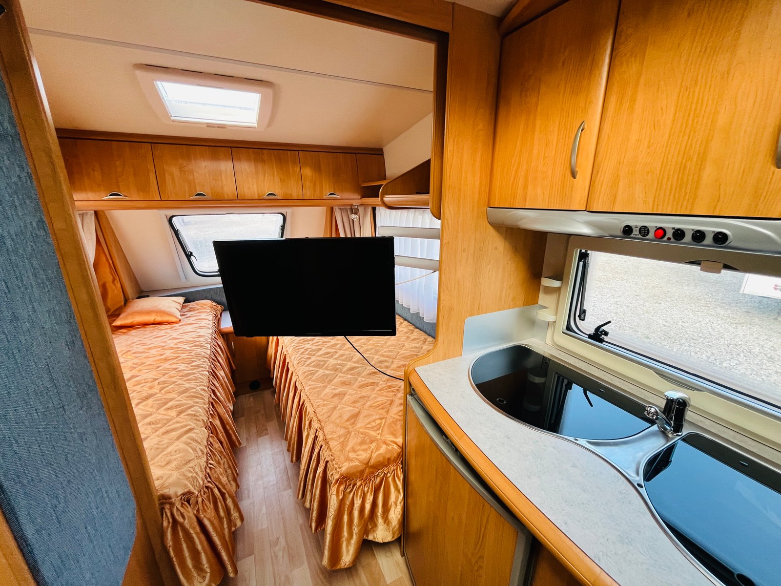 Obrázek interiéru karavanu s dvojlůžkem a kuchyňským koutem v příjemném dřevěném designu.