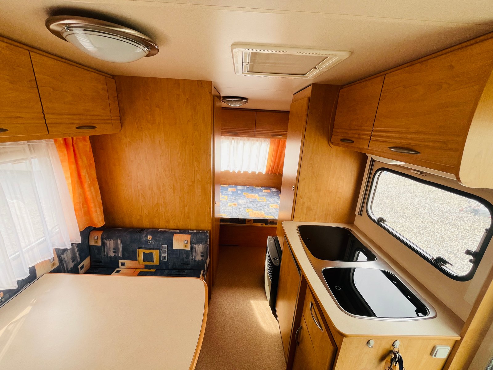 Obrázek interiéru karavanu s kuchyňským koutem a ložnicí. Vybavení v přírodním dřevě a barevnými závěsy.