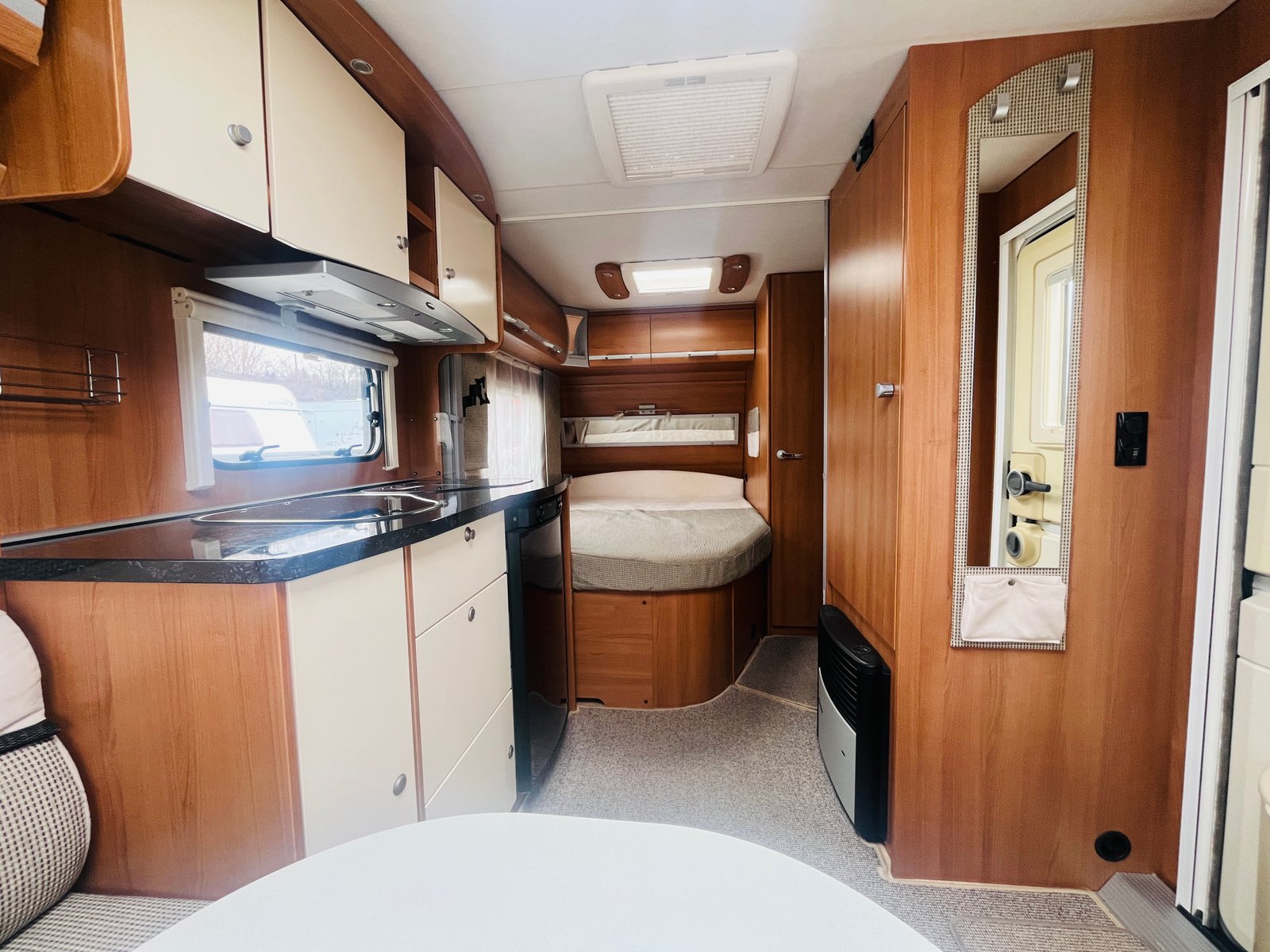 Obrázek interiéru karavanu, světlý dřevěný design, kuchyně a ložnice, pohodlné uspořádání.