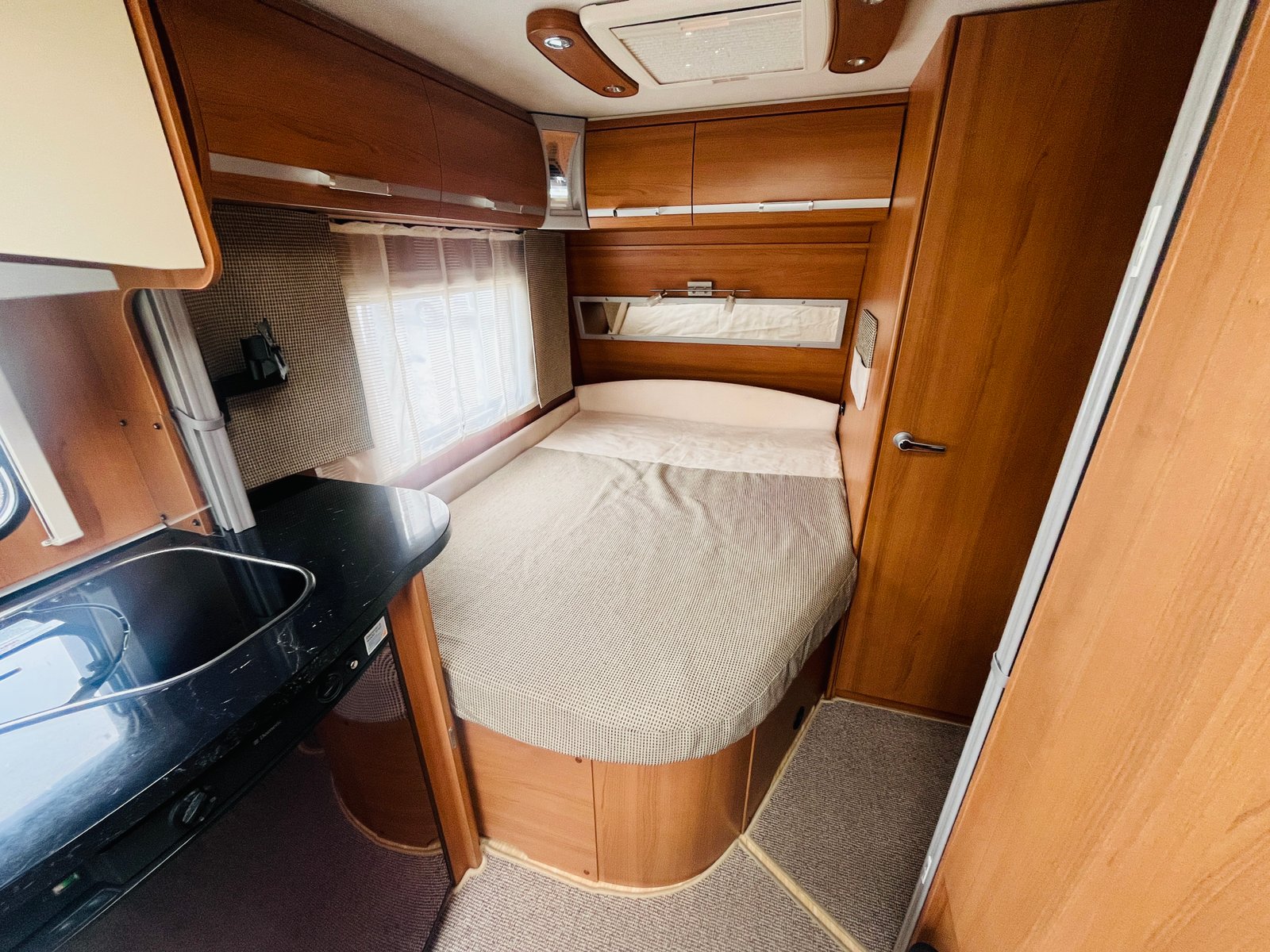 Obrázek interiéru obytného auta, útulná ložnice s dřevěným nábytkem a kuchyňským koutem.