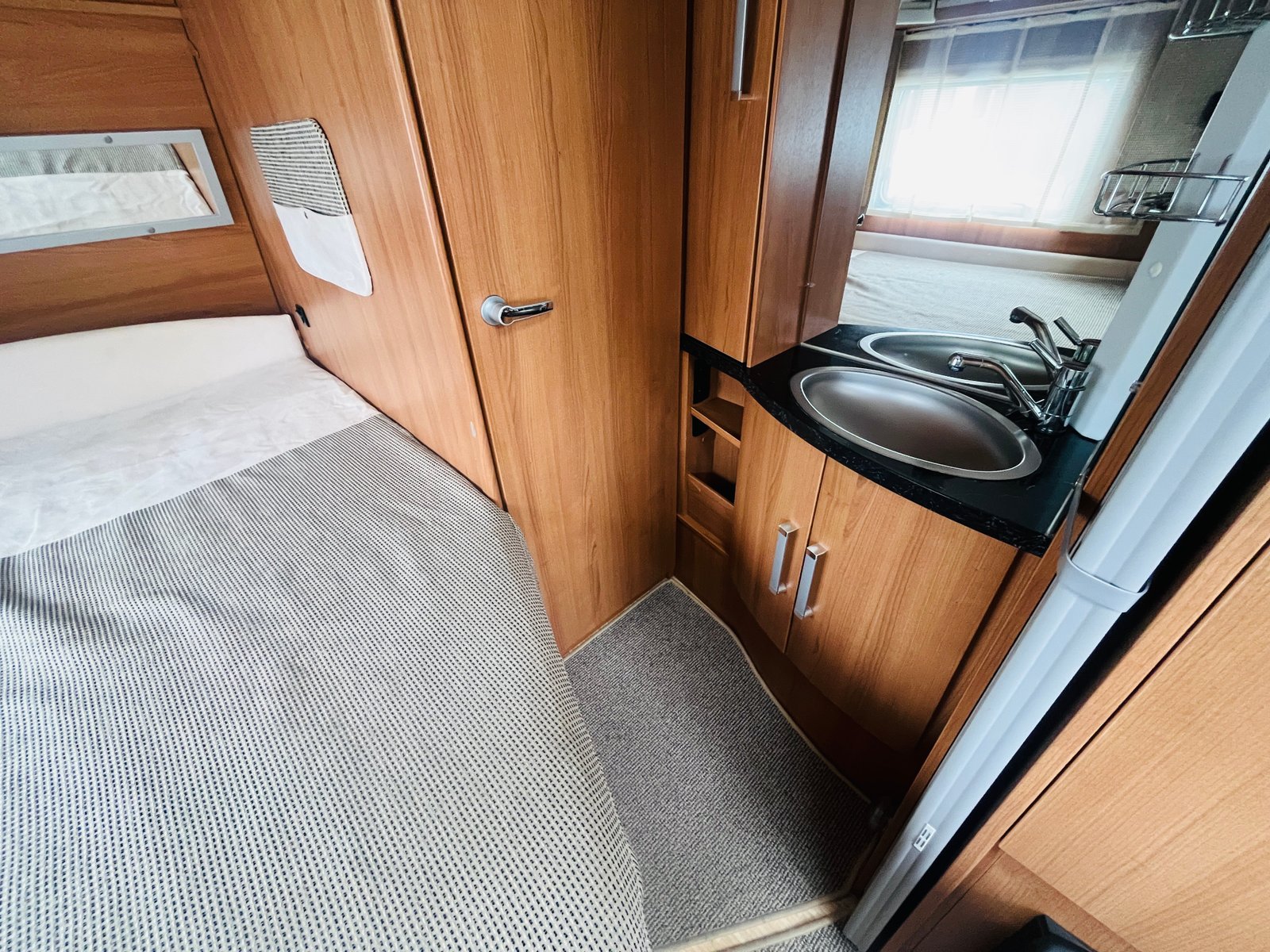 Obrázek interiéru karavanu s útulnou ložnicí a kuchyňským koutem. Moderní dřevěné provedení.