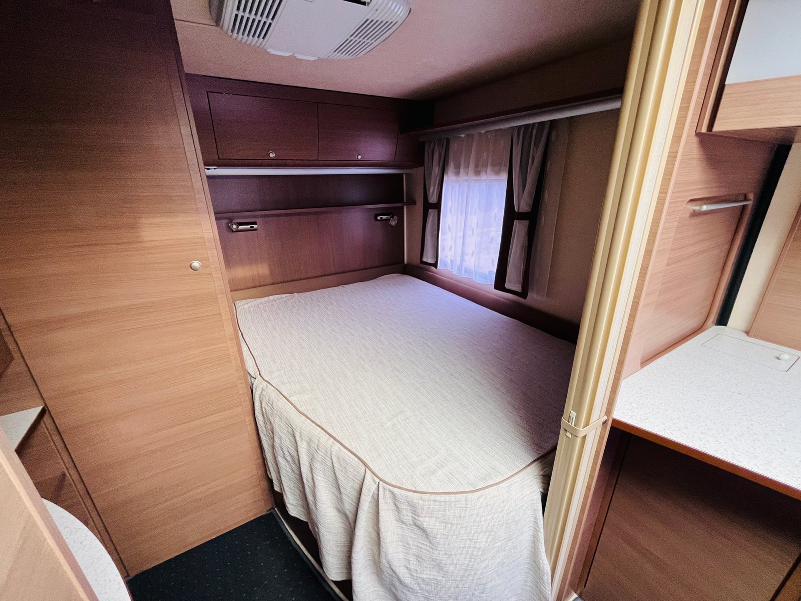 Obrázek zobrazuje útulnou ložnici v karavanu. Je zde komfortní postel a dřevěné skříňky.