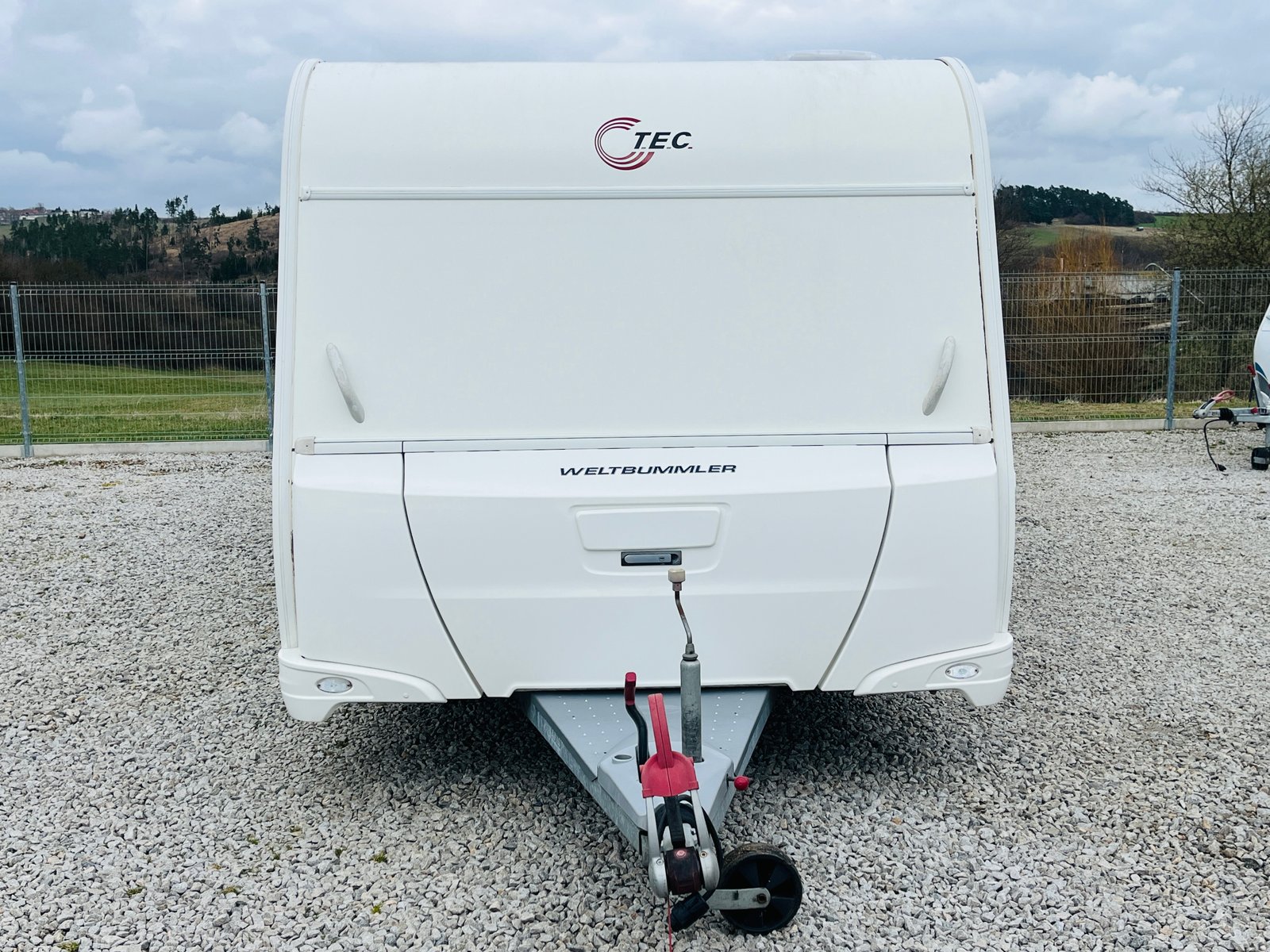 Karavan TEC Weltbummler, bílé barvy, parkovaný na štěrku v přírodě. Ideální pro dobrodružství na cestách.