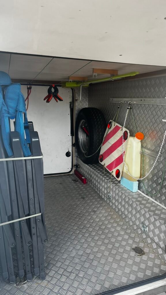 Obrázek zobrazuje úložný prostor karavanu s vybavením a náhradním kolem, což ukazuje na praktičnost pro cestování.