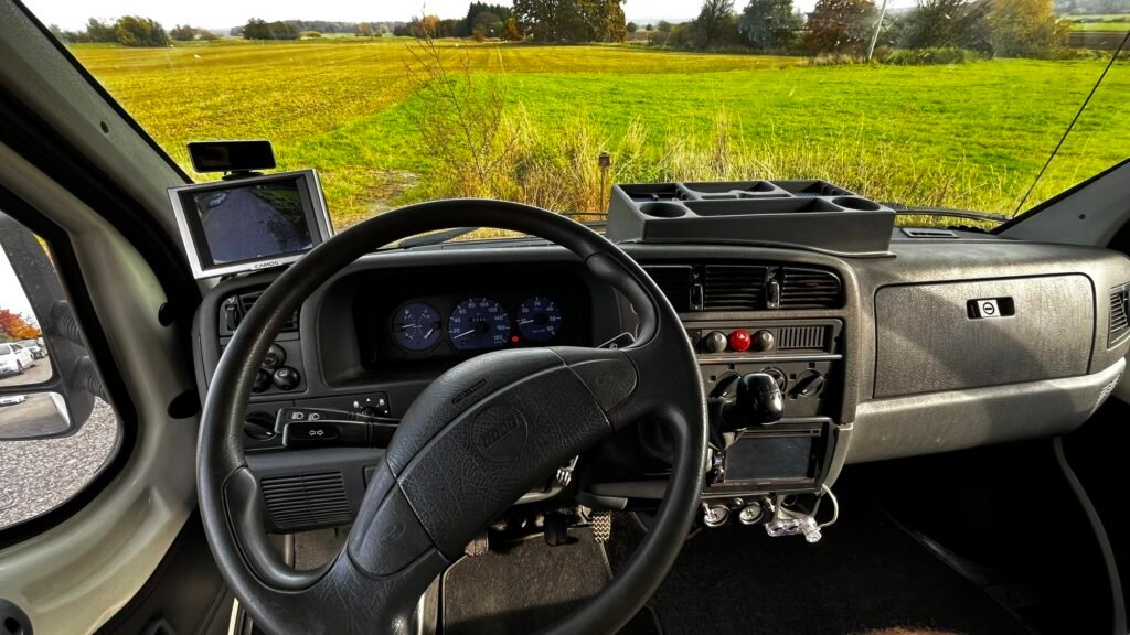 Interiér obytného auta s přehlednou palubní deskou a výhledem na zelené pole.