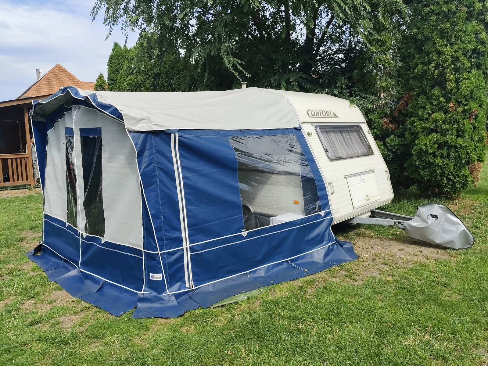 Karavan Confort je umístěn na travnaté ploše, obklopen zelení, ideální pro kempování a relaxaci.