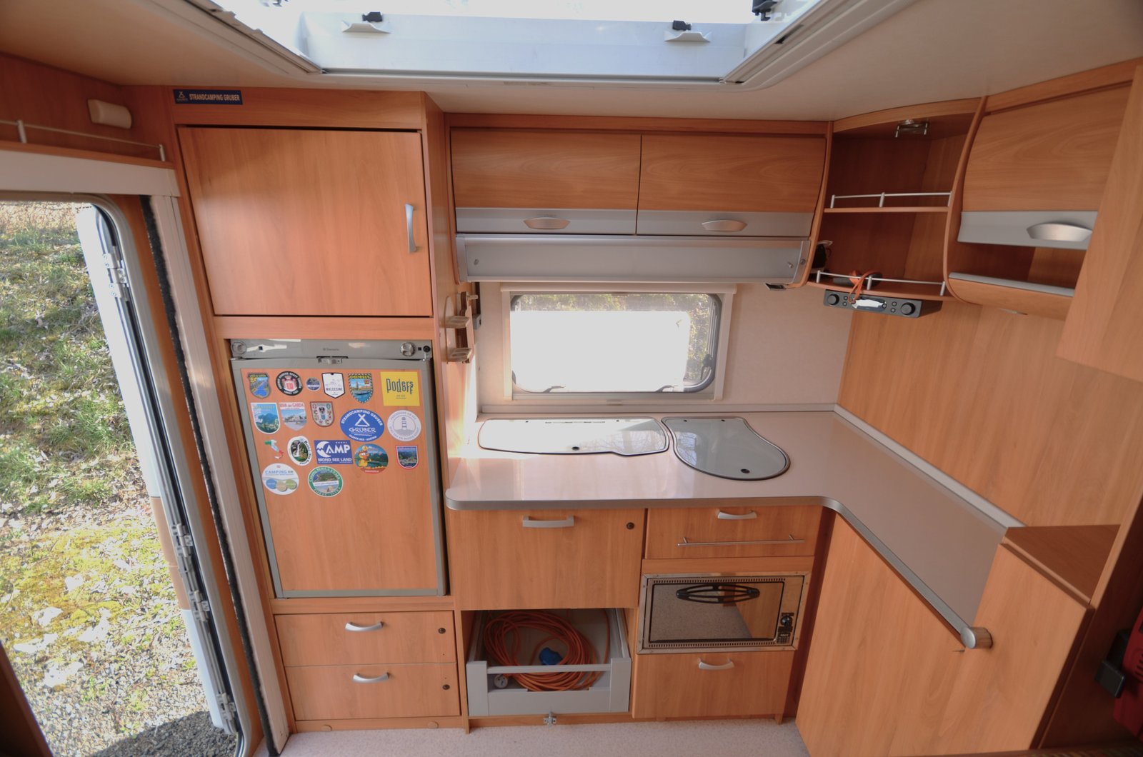 Kuchyňka v karavanu s dřezem, lednicí a úložnými prostory. Příjemné dřevěné detaily pro pohodlné vaření na cestách.