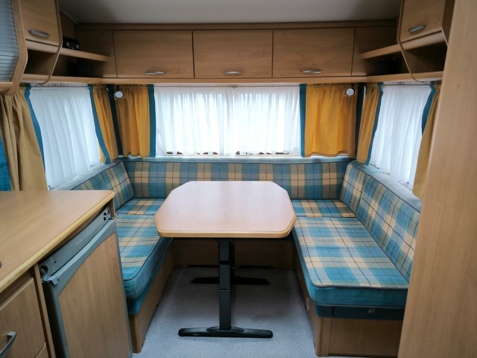 Interiér karavanu s obývacím prostorem, modro-žluté koberce a stůl pro příjemné posezení. Ideální pro dovolenou.