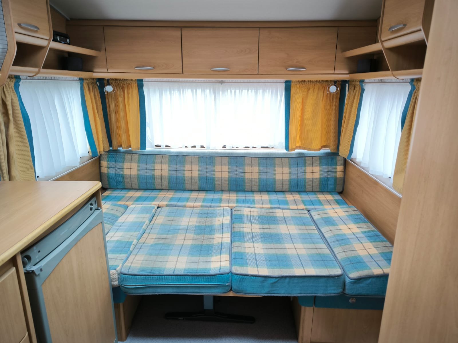 Interiér karavanu s modrými a žlutými kostkovanými sedáky a dřevěnými skříňkami. Světlo a útulná atmosféra.