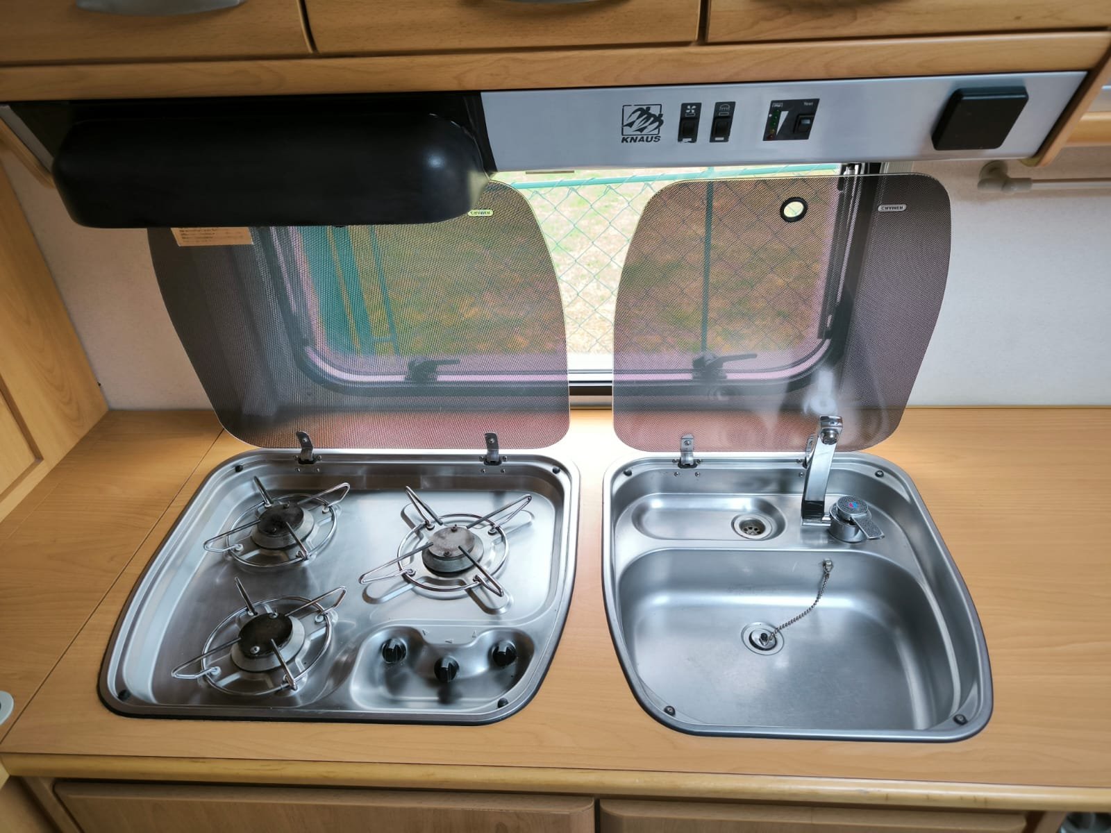Obrázek ukazuje vybavenou kuchyňku v obytném autě Knaus s trojím plynovým vařičem a dřezem.