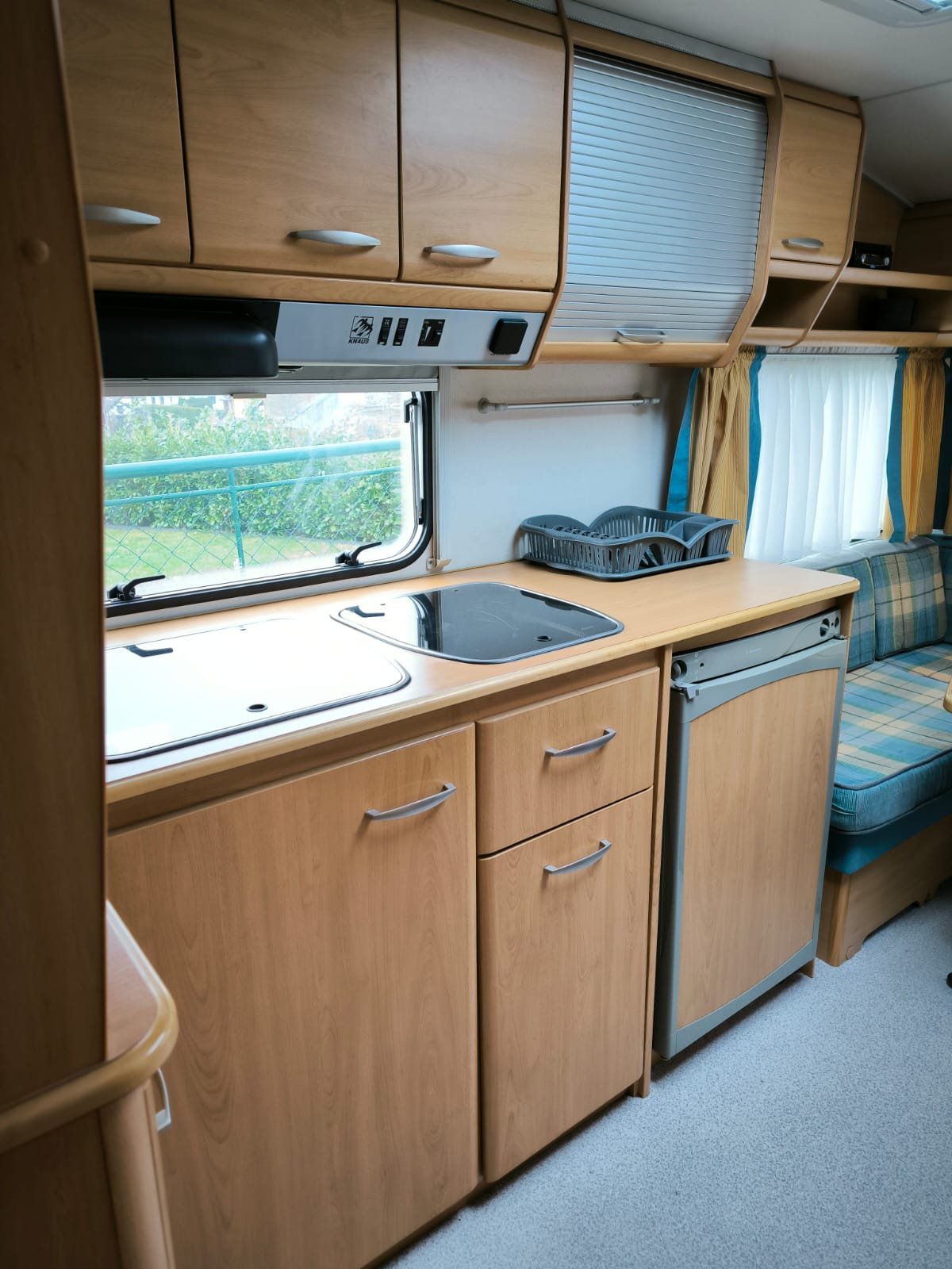 Obrázek interiéru karavanu s kuchyňskou linkou, dřezem a lednicí. Vybavení dřevěného stylu, pohodlné posezení.
