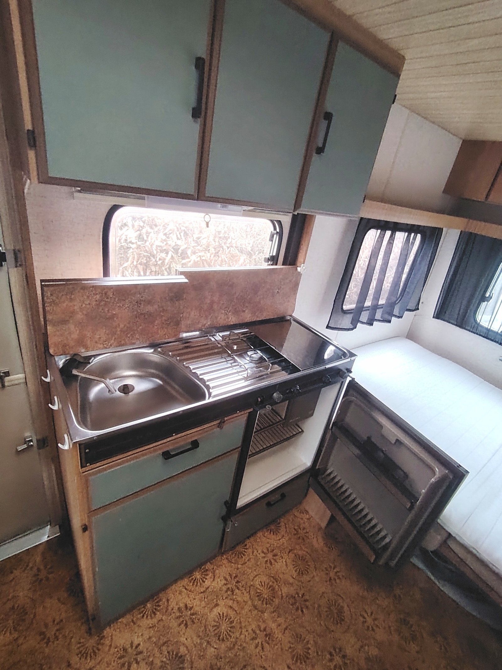 Obrázek zobrazuje interiér karavanu s kuchyňským koutem a lednicí. Praktické zařízení pro pohodlné cestování.