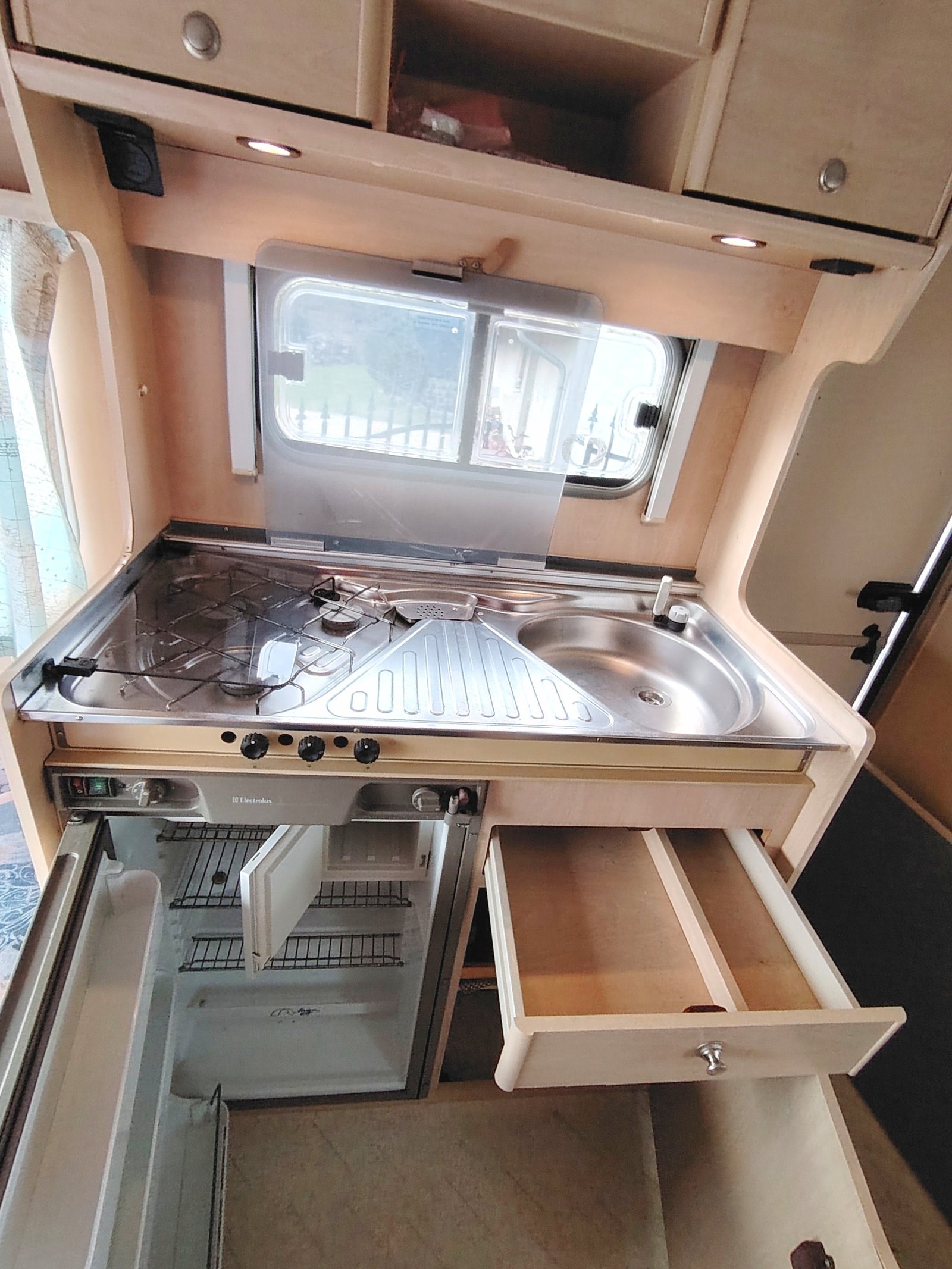 Obrázek interiéru karavanu s kuchyňským koutem, včetně varné desky a lednice. Komfortní a funkční prostor.