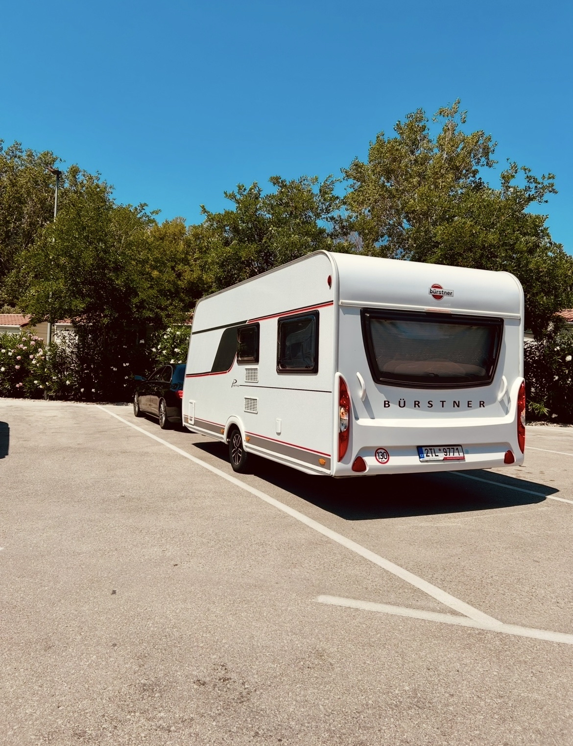 Karavan Bürstner zaparkovaný na slunném místě, obklopený zelení, ideální pro letní dobrodružství.