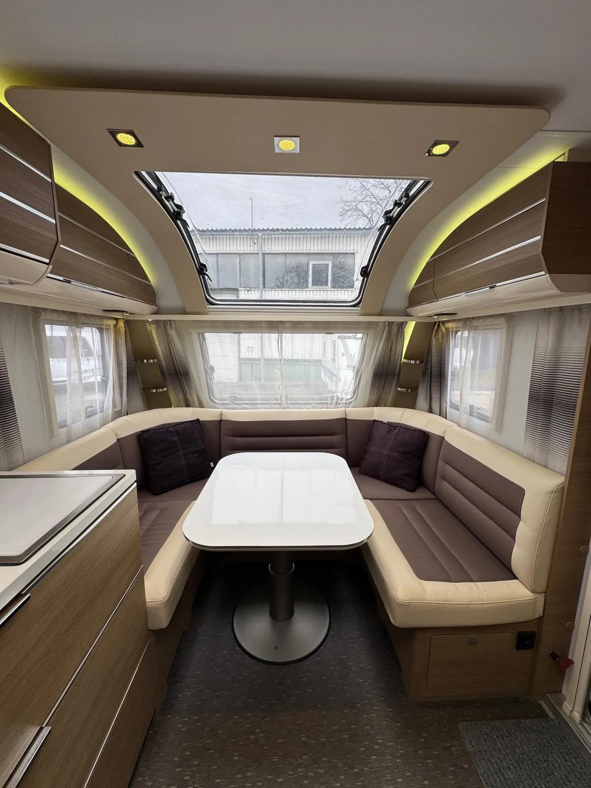 Obrázek interiéru karavanu s útulným sezením, stolem a velkým střešním oknem. Ideální pro pohodové cestování.