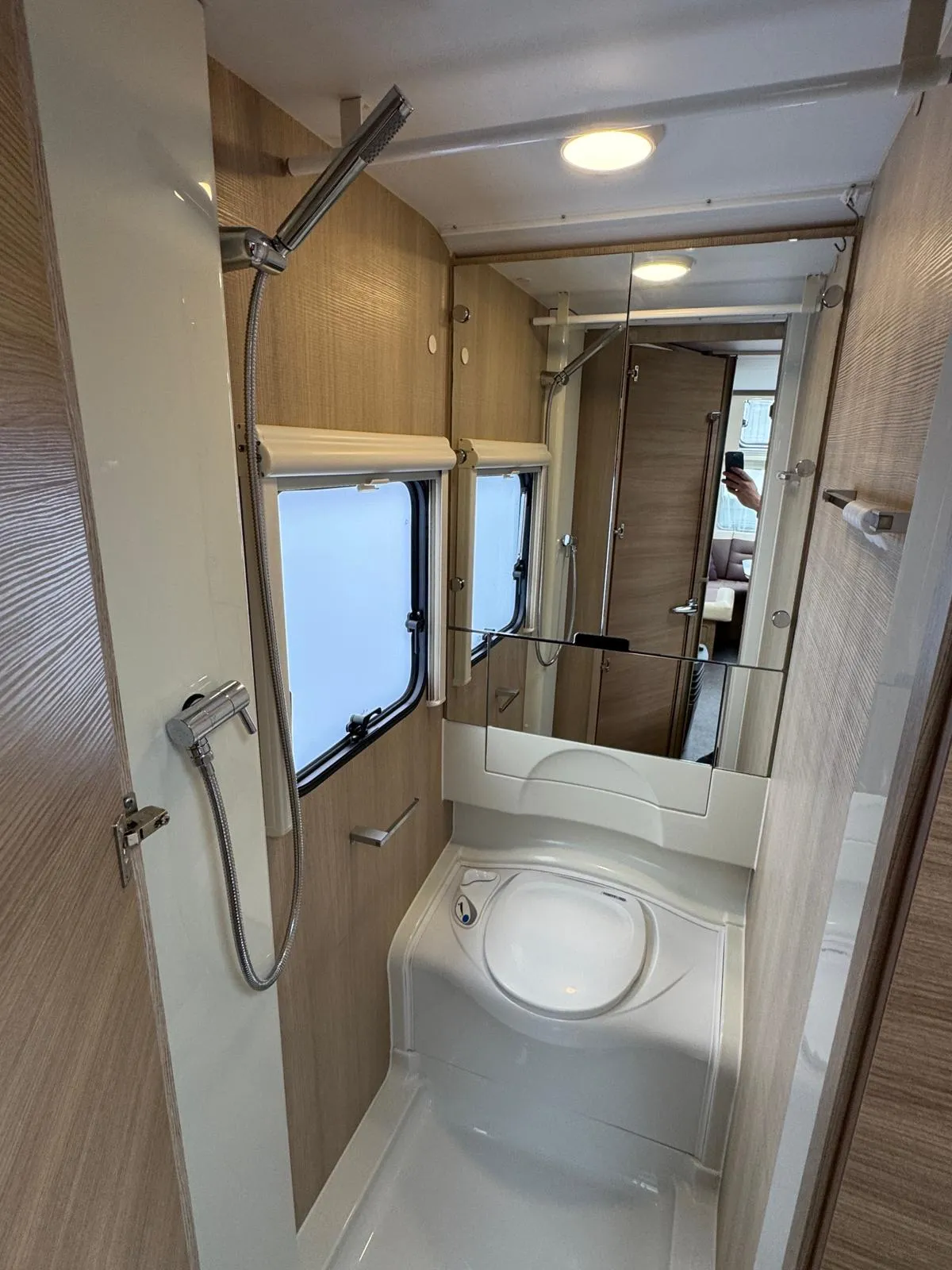 Koupelna v moderním obytném autě, zahrnující sprchu, zrcadlo a úložné prostory. Elegantní design s dřevěnými prvky.