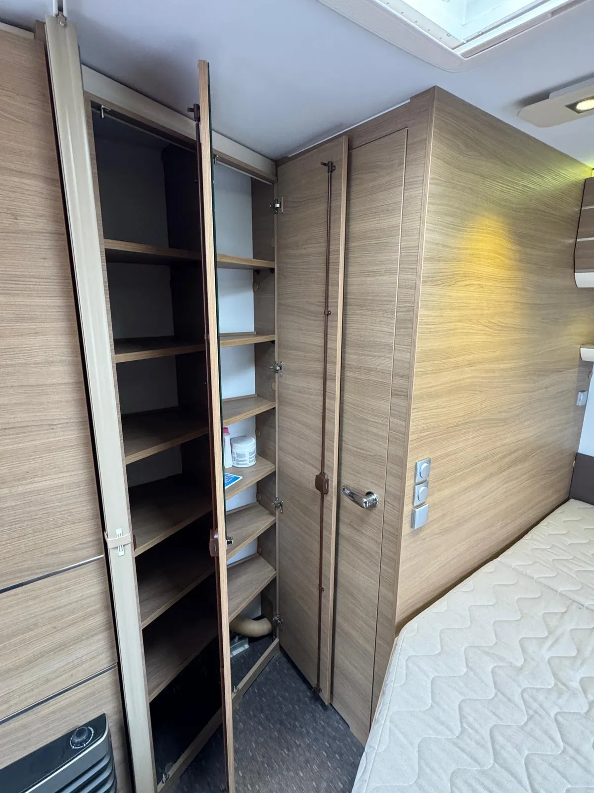 Obrázek interiéru obytného auta s úložným prostorem a manželskou postelí, moderní dřevěné prvky.