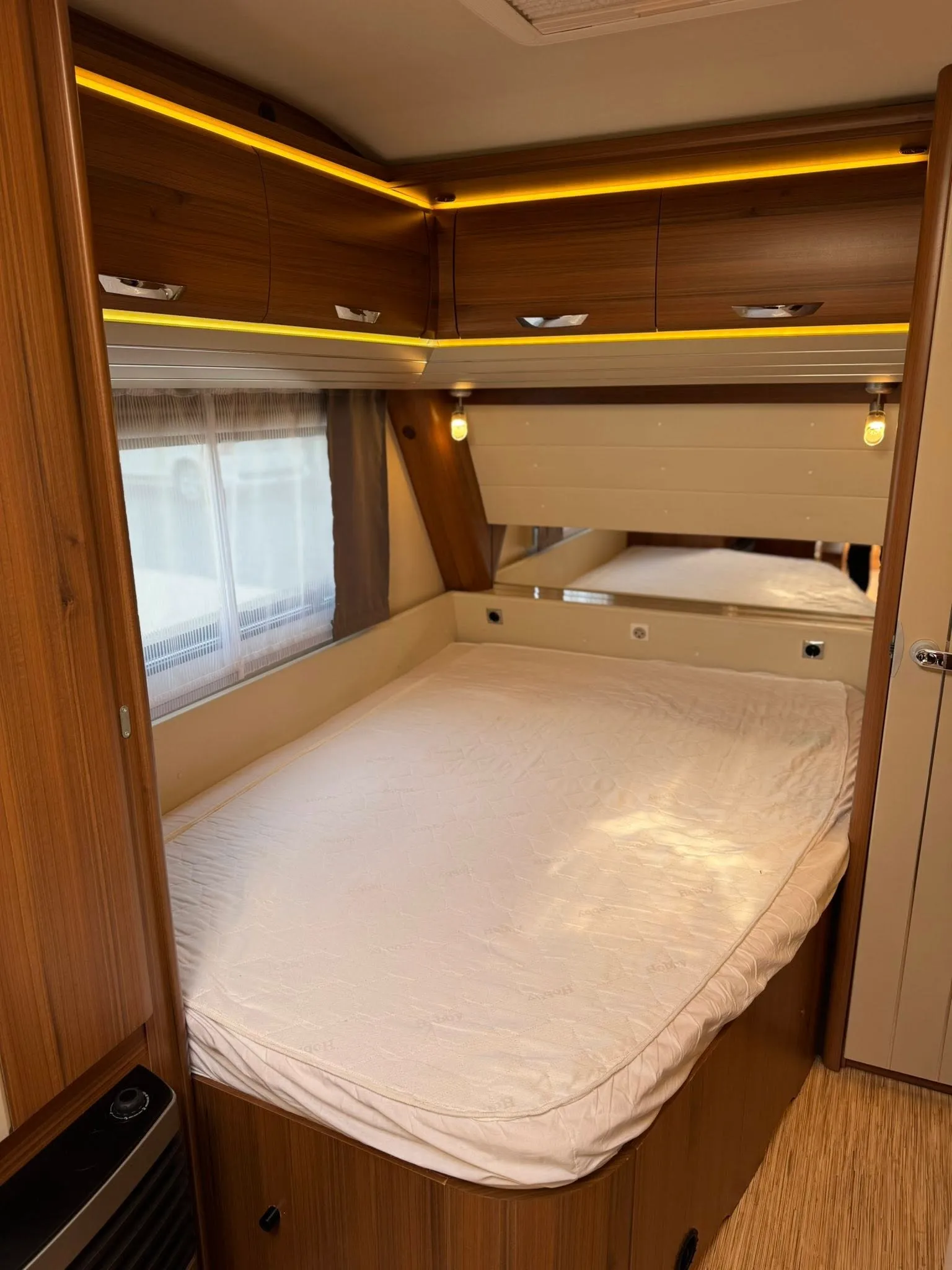 Obrázek interiéru karavanu s manželskou postelí a úložnými prostory. Moderní a útulný design pro pohodlné cestování.