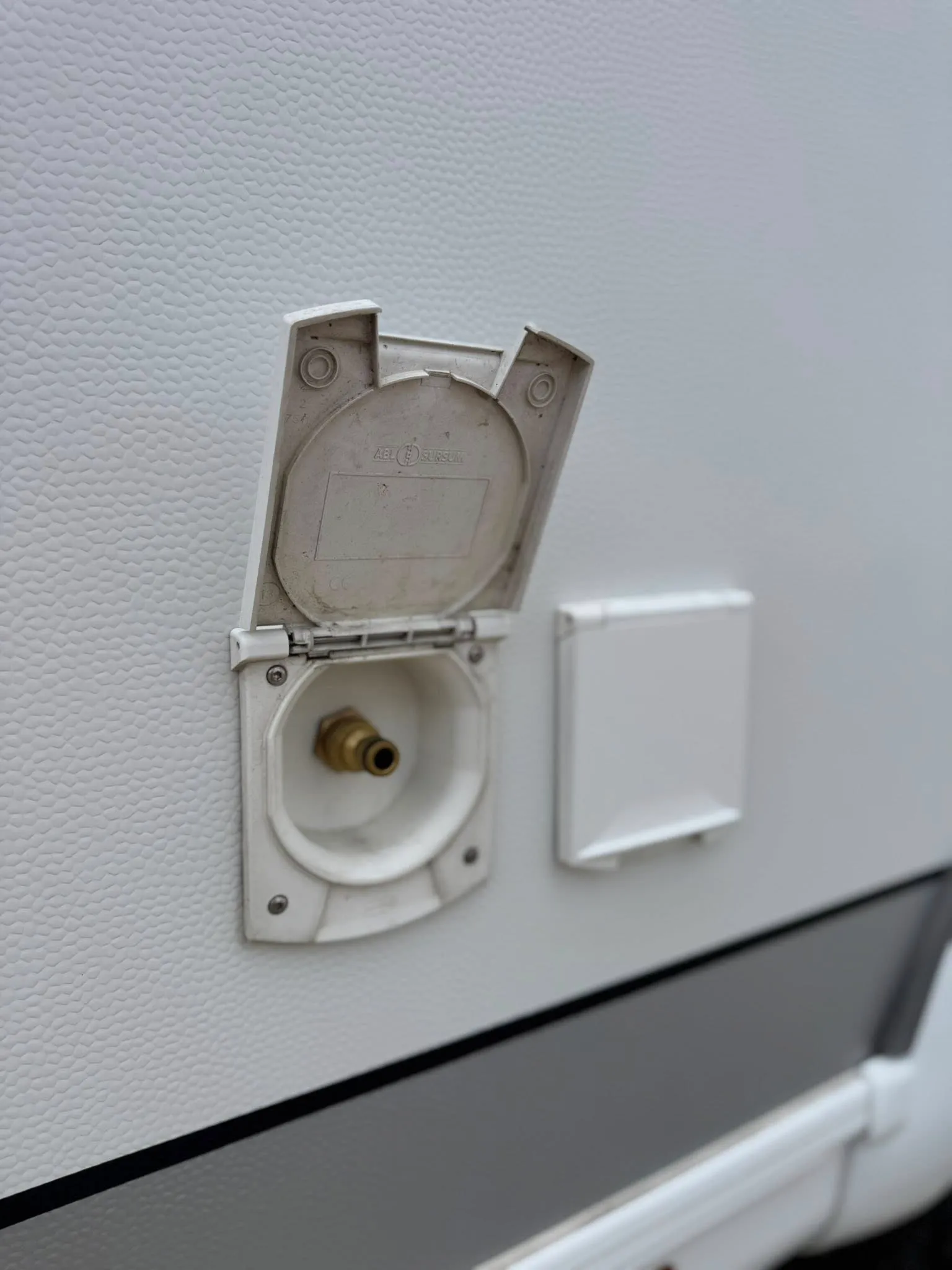 Obrázek zobrazuje vodní přípojku na karavanu s bílým exteriérem.