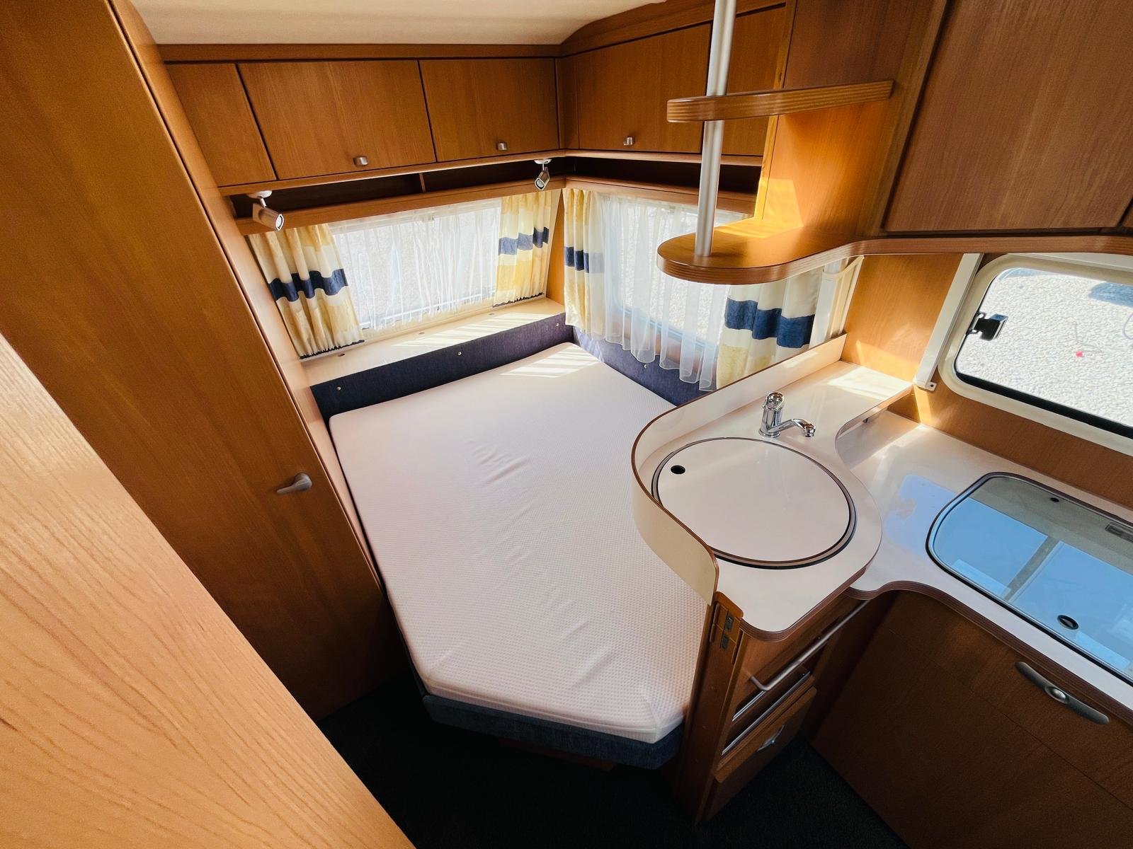 Obrázek zobrazuje interiér karavanu s manželskou postelí a kuchyňským koutem. Prostorný a útulný.