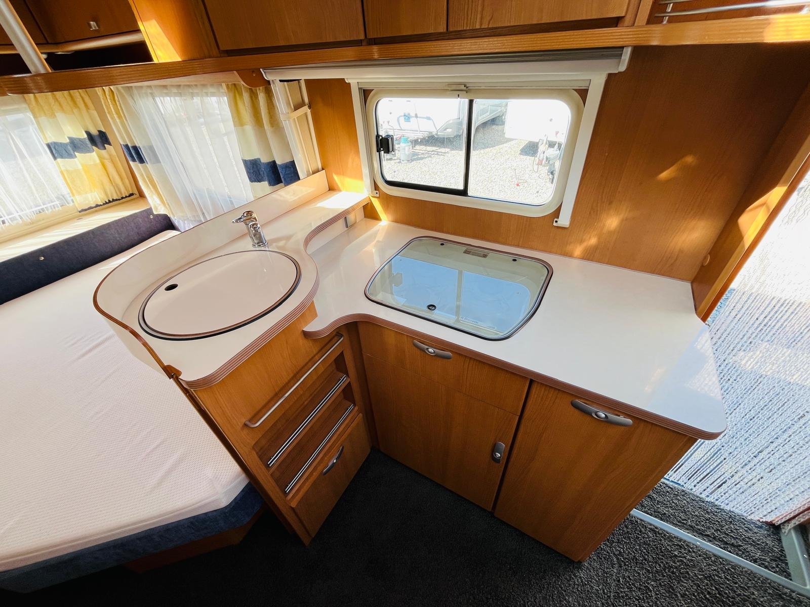 Obrázek zobrazuje interiér karavanu s kuchyňským koutem a lůžkem. Dobré uspořádání a světlý prostor.