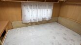 Interiér karavanu s manželskou postelí a úložným prostorem. Světlý dřevěný nábytek a záclona. Ideální pro pohodlný spánek.