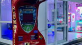 Na obrázku je herní automat Combo Shooter. Je umístěn před zábavním centrem s barevnými světly.