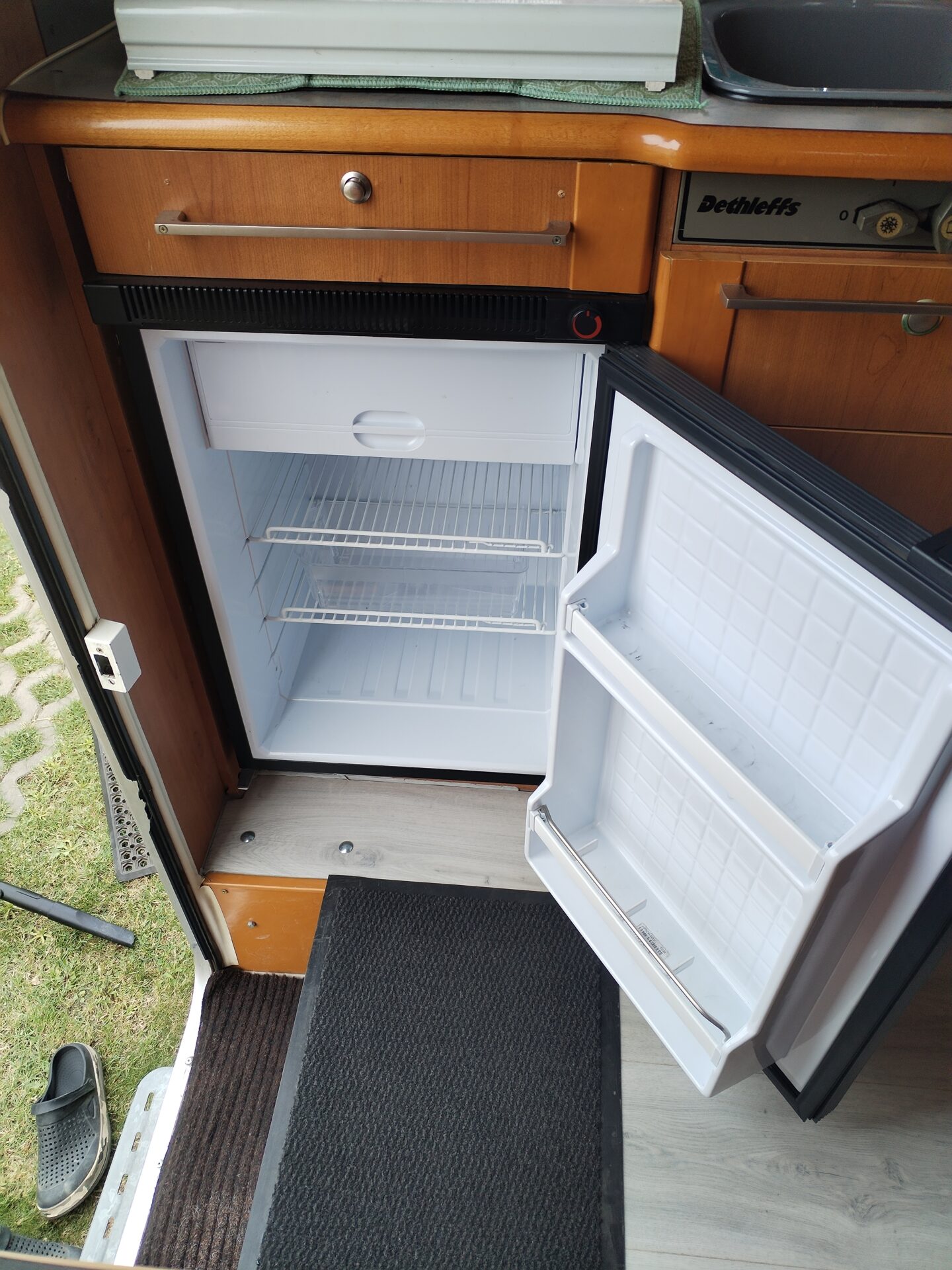 Interiér karavanu Dethleffs s otevřeným ledničkou a dřezem. Moderní design a praktické uspořádání pro pohodlné kempování.