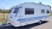 Karavan Hobby De Luxe v bílé barvě s modrými pruhy, ideální pro pohodlné cestování a kempování.