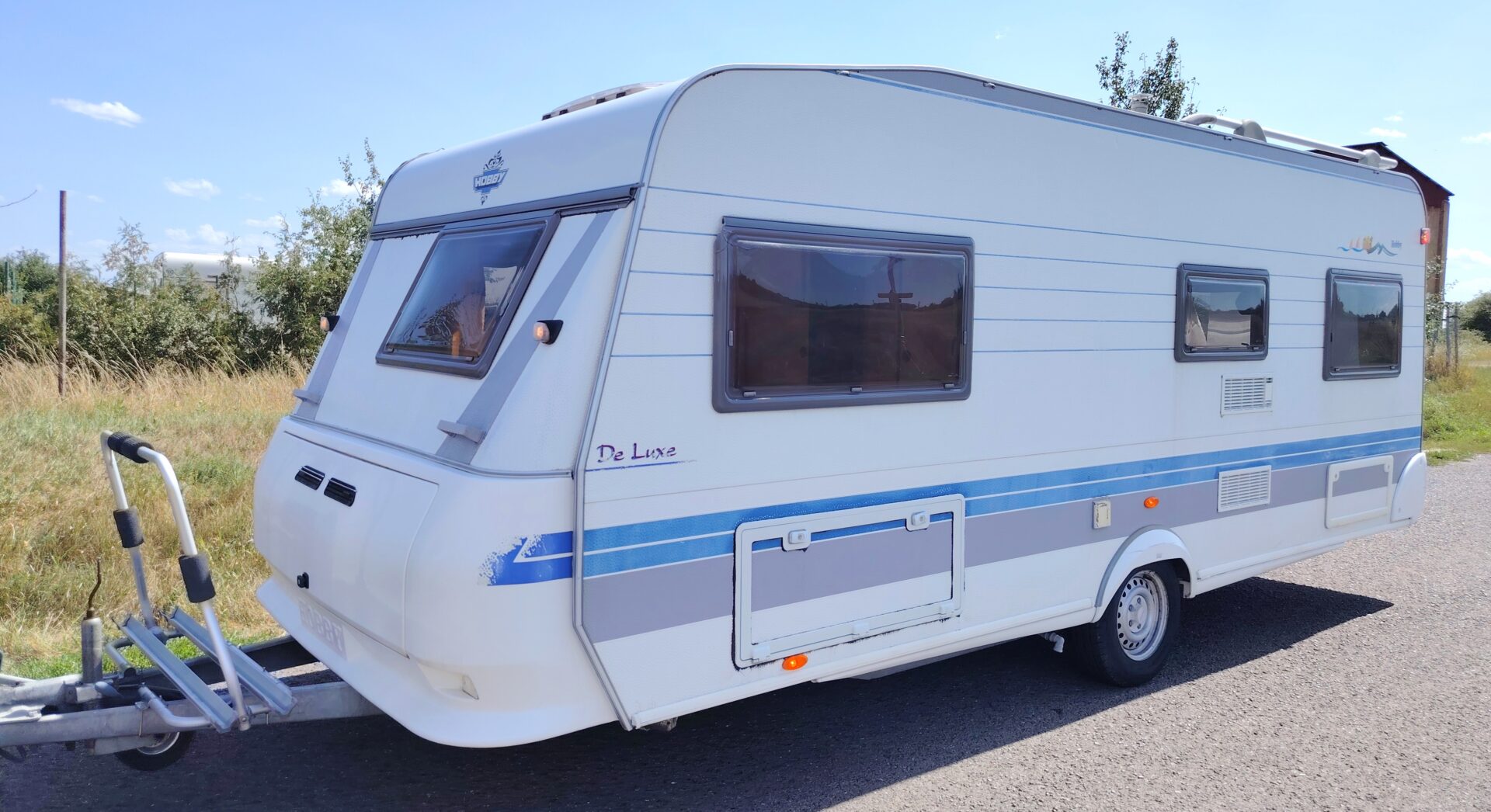 Karavan Hobby De Luxe v bílé barvě s modrými pruhy, ideální pro pohodlné cestování a kempování.