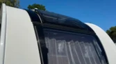 Na obrázku je karavan značky Adria. Uvnitř jsou vidět závěsy a prosklená střecha.